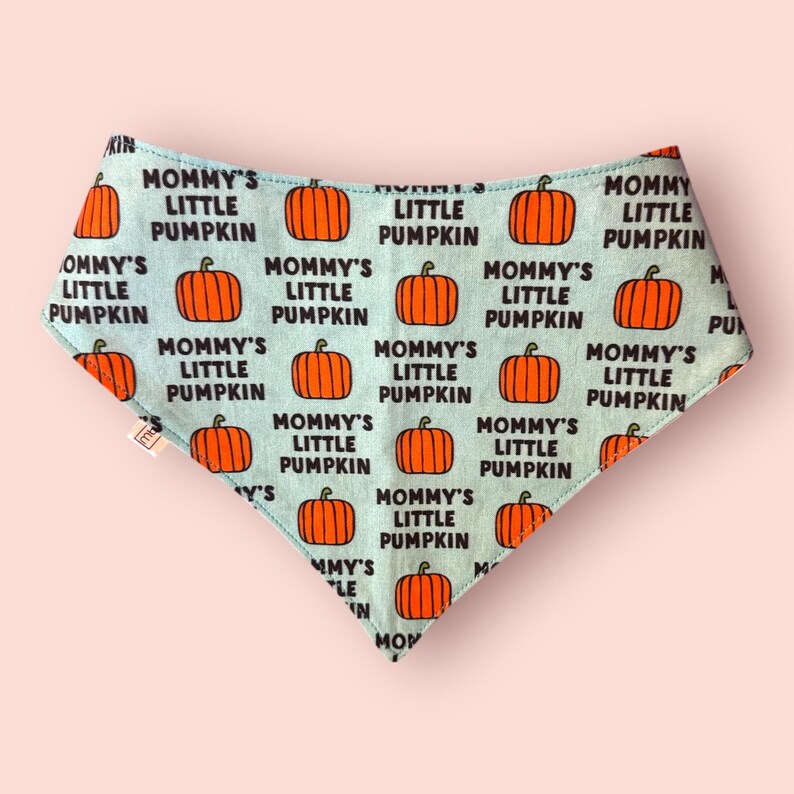 May include: A triangular bandana with a light blue background featuring a repeating pattern of orange pumpkins and the black text Mommy's Little Pumpkin. The fabric has a small white tag on the left side and a pointed bottom edge.