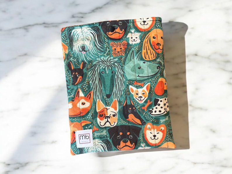 May include: A rectangular quilted fabric sleeve featuring a repeating pattern of various dog breeds on a dark teal background. The sleeve includes a small white rectangular label with the black text mb printed on the bottom left corner.