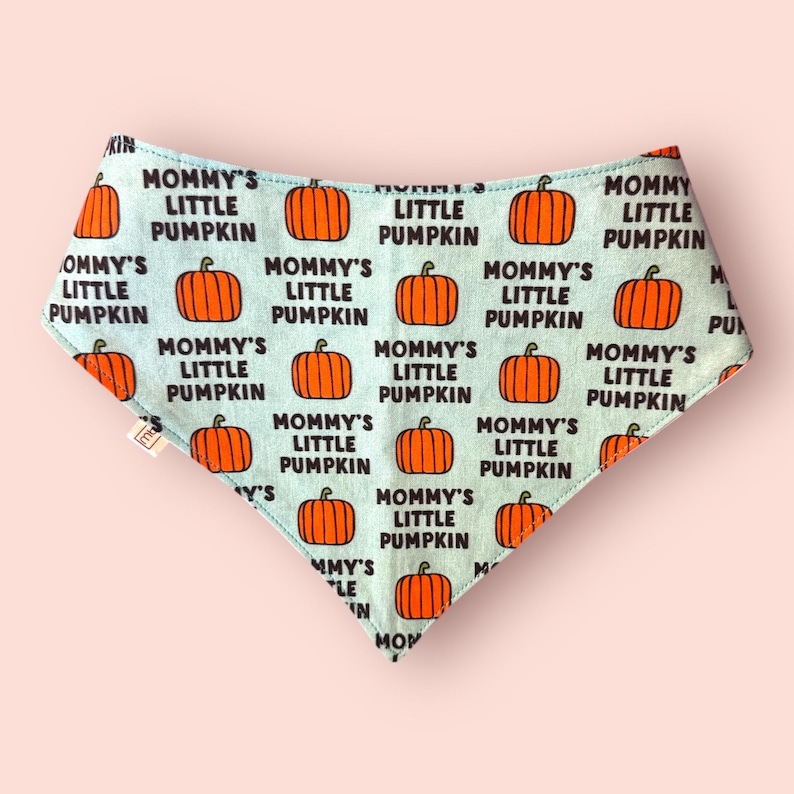 May include: A triangular pet bandana featuring a light blue background with a repeating pattern of orange pumpkins and the black text Mommy's Little Pumpkin. The fabric has a small white tag on the left side and a pointed bottom edge.