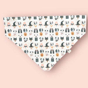 May include: A triangular pet bandana featuring a repeating pattern of various dog breeds dressed in Halloween costumes, including witch hats, masks, and glasses, alongside orange jack-o'-lanterns and black trick-or-treat bags on a white background.