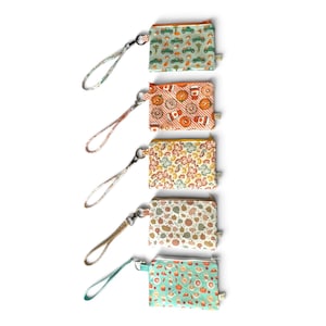May include: Five rectangular zippered pouches with wrist straps, each featuring a distinct seasonal pattern: green trucks, orange donuts and coffee, autumn leaves, assorted pumpkins, and a light blue print with various small fall-themed icons.