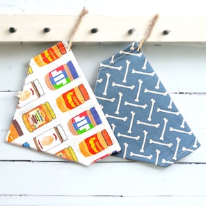 May include: Two dog bandanas hanging on a white wooden rack. One bandana is white with a peanut butter jar pattern and the other is blue with a white bone pattern.