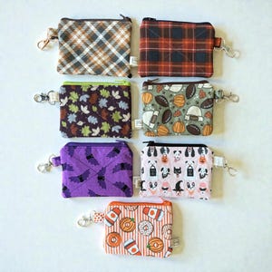 May include: A selection of small, rectangular fabric pouches with zippers and key rings. The pouches display diverse patterns, including plaid, leaves, footballs, bats, dogs, and donuts. Each pouch has a unique colour scheme.