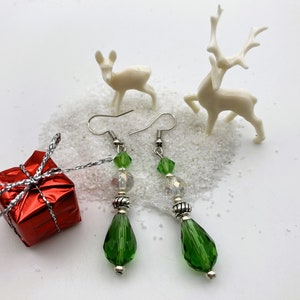 May include: A pair of dangle earrings with green teardrop-shaped beads, clear beads, and silver accents. The earrings are displayed with a small red gift box and miniature white reindeer figurines on a white, snowy surface.