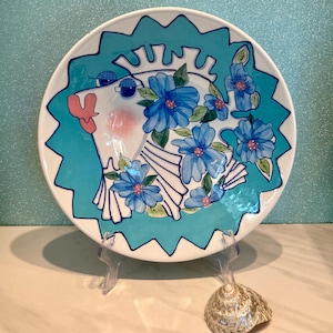 May include: A decorative ceramic plate featuring a whimsical fish design. The fish is white with blue accents, adorned with blue floral patterns. The plate has a turquoise border and is displayed on a clear stand, with a seashell nearby.