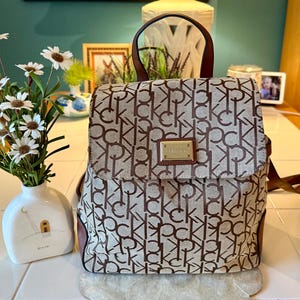 May include: A beige and brown backpack featuring a repeating CK monogram pattern, a top handle, and a rectangular gold-toned metal plaque on the front flap. The bag has brown leather-like trim and adjustable straps, with a structured silhouette.