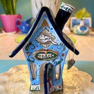May include: A ceramic house figurine with a blue roof and walls, featuring a chimney and an open door. The house is decorated with floral and bird motifs, and the word "Peace" is displayed on the front.