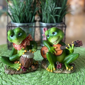 May include: Two green frog figurines. One frog plays a drum, wearing a red and white polka dot scarf. The other frog plays a small guitar. Both frogs sit on a base with flowers and leaves. The background includes green plants.