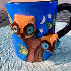 May include: A blue ceramic mug with a black handle, featuring a 3D owl design. The mug has a night sky scene with a moon and stars. The owls are brown and orange with blue eyes. The mug sits on a light blue woven mat.