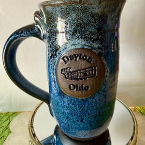 May include: A handcrafted ceramic mug with a blue and black speckled glaze. The mug features a large handle and a circular emblem with the words "Dayton Ohio" embossed on it. The mug is displayed on a decorative gold-colored stand.
