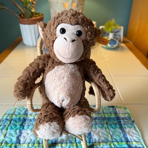 May include: A plush brown monkey with a light brown face, belly, and feet sits on a small wicker chair. The monkey has large black eyes and is positioned on a blue and green plaid quilted mat. The background includes a plant in a white pot.