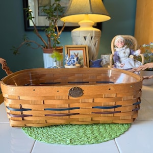 May include: A rectangular woven basket with brown leather handles. The basket is made of light brown wood with accents of blue and brown woven through the sides. A small metal emblem is attached to the front.