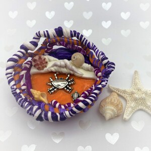 May include: A handcrafted, miniature beach scene with a vibrant orange base, surrounded by twisted purple, white, and orange clay. Adorned with seashells, a starfish, and a crab charm. A small turtle charm is also present. The scene evokes a coastal theme.