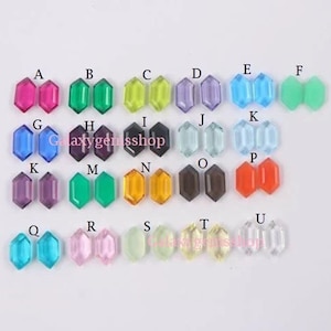 Op de afbeelding: Assortiment van 20 paar kleine, gefacetteerde, achthoekige glazen edelstenen in verschillende kleuren, waaronder roze, paars, blauw, groen, geel, oranje en helder. De edelstenen zijn gerangschikt in rijen die zijn gelabeld met de letters A tot en met U.