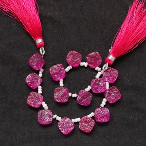 May include: A strand of pink gemstone beads with a square shape and a frosted finish. The beads are strung on a white cord and have pink tassels on each end.