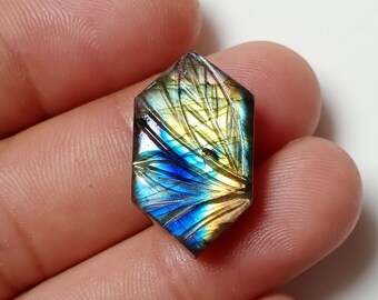 May include: A faceted, hexagonal gemstone with intricate carved details. The stone displays a vibrant play of color, with hues of blue, yellow, and green. The gemstone is held in a hand, showcasing its size and shape.