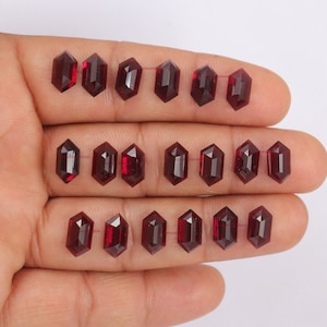 May include: A selection of dark red, hexagonal gemstones, each with multiple facets. The gems are presented against a plain backdrop, highlighting their polished surfaces and clarity. The gemstones are approximately 1.3 cm in length.
