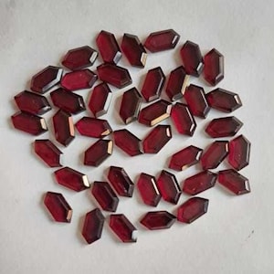 May include: A collection of dark red, hexagonal gemstones scattered on a white surface. The faceted stones display a deep, rich colour and polished surfaces. These could be used for jewellery making or other crafts.