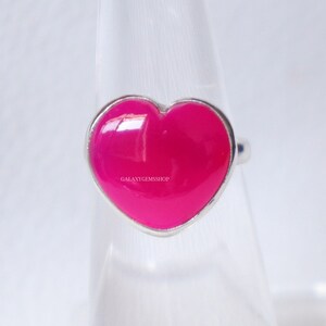 May include: A silver ring featuring a vibrant, hot pink heart-shaped stone. The ring has a polished finish and the text "GALAXYGEMSSHOP" is visible on the stone. The ring is displayed on a clear stand.