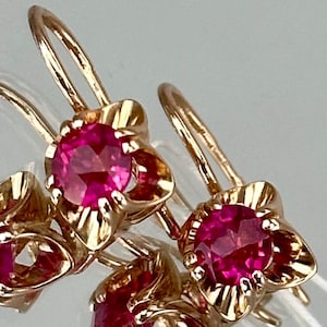May include: Gold-tone earrings with bright pink gemstones. Each earring features a flower-shaped setting with a round, faceted gemstone at the center. The earrings have a classic hook design.