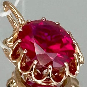 May include: A gold pendant with a large, oval-cut ruby gemstone. The ruby is a deep red hue and is set in an ornate gold setting. The pendant has a loop for a chain, and is reflected in the surface below.