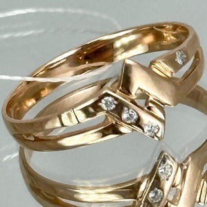 May include: Two gold rings with a V-shaped design, each adorned with small, sparkling diamonds. The rings are displayed on a reflective surface, showcasing their intricate details and warm, golden hue. The rings are a classic jewelry item.