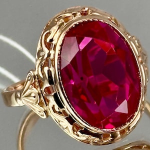 May include: A gold ring with an oval, faceted, ruby-red gemstone. The ring has an ornate, filigree design around the gemstone and on the band. The ring is set against a reflective surface.