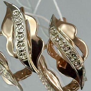May include: A pair of gold-coloured earrings with a leaf design. Each earring has a central row of small, sparkling stones. The earrings have a polished finish and a secure clasp.
