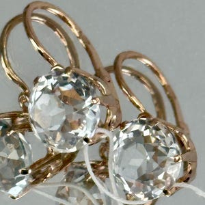 May include: Close-up of gold-colored earrings with large, clear, faceted gemstones. The earrings have a lever back closure. The gemstones are round and appear to be set in a gold-colored metal. The earrings are displayed against a light background.