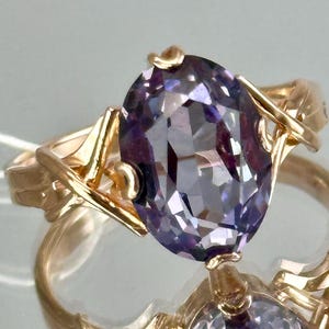 May include: A gold ring featuring a large, oval-cut, purple gemstone. The ring's band and setting are also gold, with an intricate, stylized design. The gemstone reflects light, showcasing its facets. The ring is displayed on a reflective surface.