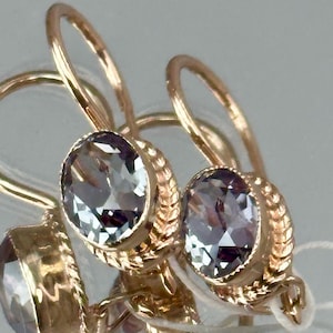 Könnte beinhalten: Ein Paar Ohrhänger aus Roségold mit oval geschliffenen, hellvioletten facettierten Steinen. Jeder Stein ist in einer strukturierten Metallfassung im Seil-Design gefasst und verfügt über einen gebogenen Haken. Glänzende metallische Oberfläche.