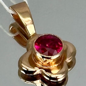 May include: A gold pendant featuring a four-petaled flower design with a central round, faceted, deep pinkish-red gemstone set in a bezel. The pendant includes a polished gold bail at the top, with a white string attached for display purposes.