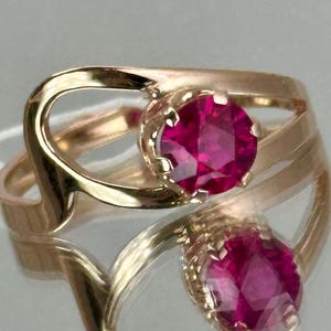 Op de afbeelding: Een gouden ring met een grote, ronde, helderrode edelsteen. De band van de ring heeft een uniek, open ontwerp met vloeiende lijnen. De edelsteen is gezet in een gedetailleerde, sierlijke zetting, die de elegante stijl van de ring benadrukt.