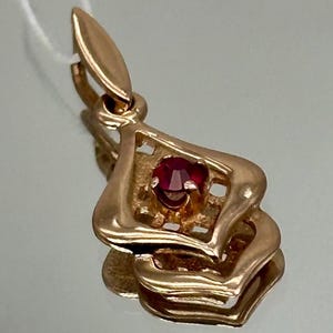 Peut inclure: Un pendentif en or avec une pierre précieuse rouge au centre. Le pendentif présente un design ajouré orné en forme de goutte d'eau. La pierre précieuse est d'un rouge profond et est sertie dans un cadre carré. Le pendentif est suspendu à une petite boucle.