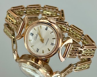 May include: A gold-tone wristwatch with a white face and black Roman numeral hour markers. The watch has a gold-tone band with a textured design. The watch face says "Chaika" and "УКРАИНА".