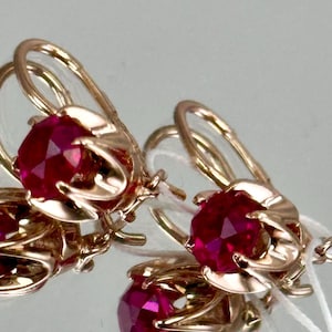 May include: Close-up of gold earrings with a floral design. Each earring features a large, faceted, deep red gemstone. The earrings have a lever-back closure. The gemstones are the focal point of the jewellery.