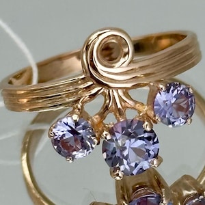 May include: A gold-colored ring with three light purple gemstones. The ring has a unique design with a swirl at the top and a textured band. The gemstones are round and set in a cluster.