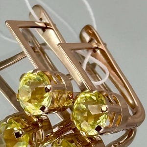 Könnte beinhalten: Nahaufnahme von goldfarbenen Ohrringen mit geometrischem Design. Jeder Ohrring hat einen quadratischen Rahmen und einen runden, gelben Edelstein. Die Ohrringe werden vor einem hellen Hintergrund präsentiert, der ihre reflektierenden Oberflächen hervorhebt.