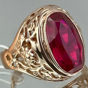 Op de afbeelding: Een gouden ring met een sierlijk ontwerp met een grote, ovale, rode edelsteen.