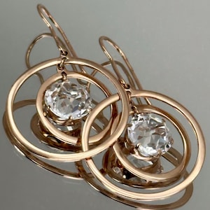 May include: Rose gold earrings featuring a large, clear, round gemstone suspended within a series of concentric circles. The earrings have a hook closure and reflect light, creating a sparkling effect.