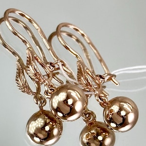 May include: Rose gold earrings featuring a ball pendant. The earrings have a lever back closure and a decorative leaf design. The ball pendant is smooth and reflective. The earrings are likely made of a precious metal.