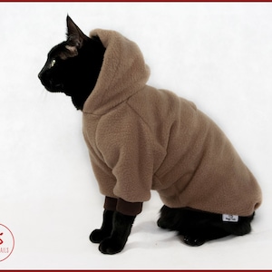以下が含まれることがあります： 茶色のフリースのフード付きセーターを着た黒い猫。セーターは茶色のフードと茶色のボディで、後ろには「Meggy Trails」と書かれた白いタグが付いています。