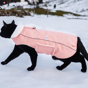 以下が含まれることがあります： ピンクのフリースの裏地が付いた反射ストリップのある犬用コートを着た黒い猫。コートはジッパーで閉じており、背面にポケットが付いています。猫は雪の上を歩いています。