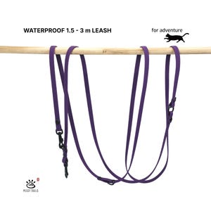 Può includere: Un guinzaglio viola impermeabile con componenti in metallo nero, recante la scritta WATERPROOF 1.5 - 3 m LEASH, for adventure, e la sagoma di un gatto. Il guinzaglio è appeso a un bastone di legno con il logo Meggy Trails nell'angolo.