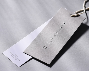 Pode incluir: Uma etiqueta de papel cinza com o texto "FASHION STYLE" em relevo. A etiqueta está presa a uma etiqueta de papel branco com o texto "CREATE ORDINARY OR NATURAL ADVOCATING".
