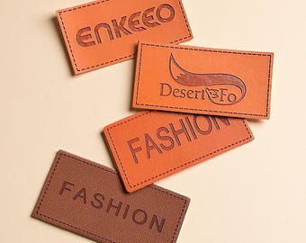 May include: Four rectangular leather labels in shades of brown and orange. The labels feature stitched edges and embossed text, including "ENKEECO", "Desert Fo", and "FASHION". The labels are likely for clothing or accessories.