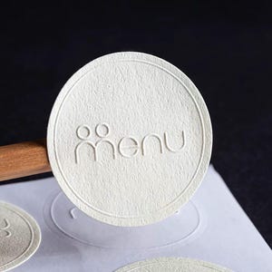 Puede incluir: Primer plano de una pegatina redonda blanquecina con la palabra "men&uacute;" en relieve en una fuente sans-serif. La pegatina tiene una superficie texturizada y un borde decorativo. La palabra "men&uacute;" est&aacute; encima de un gr&aacute;fico estilizado de dos personas.