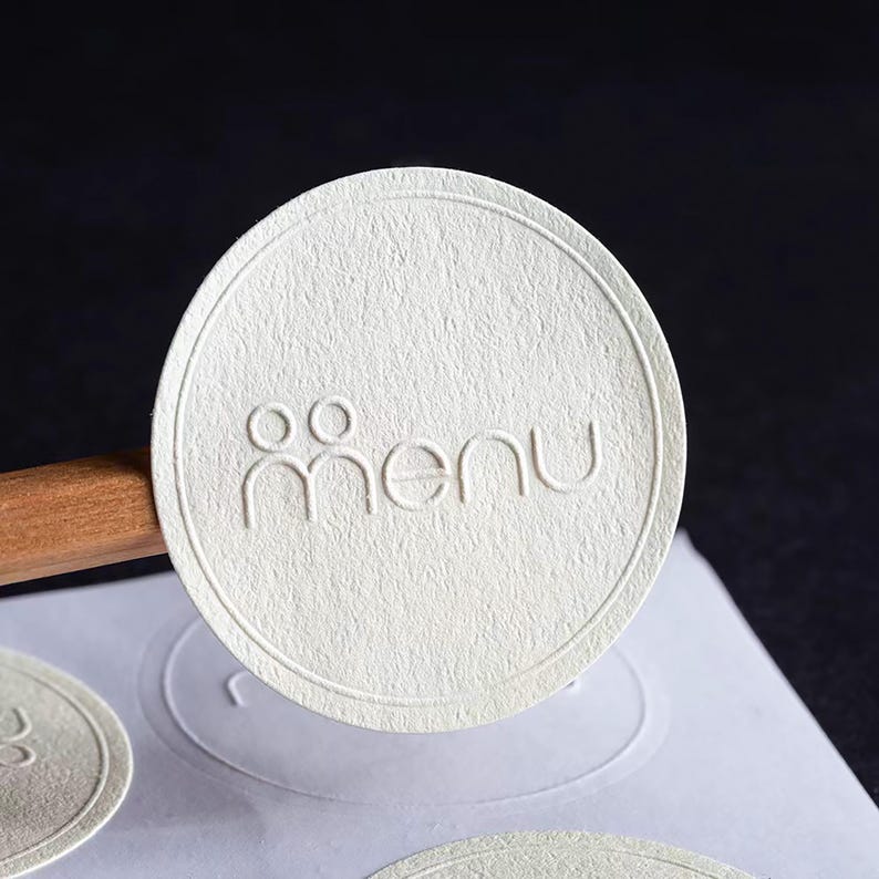 Puede incluir: Primer plano de una pegatina redonda blanquecina con la palabra "men&uacute;" en relieve en una fuente sans-serif. La pegatina tiene una superficie texturizada y un borde decorativo. La palabra "men&uacute;" est&aacute; encima de un gr&aacute;fico estilizado de dos personas.