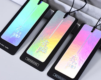 May include: Three iridescent holographic hang tags with black backing. Each tag features a different color gradient and the text "She is enjoying" printed in white on the black backing. The text "本品" is printed in white on the iridescent holographic surface.