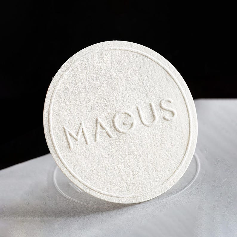 Puede incluir: Una etiqueta redonda de papel blanquecino con la palabra "MAGUS" en relieve. La etiqueta tiene una superficie texturizada y un borde fino en relieve. El fondo es blanco y negro, con un enfoque suave. Ideal para branding.